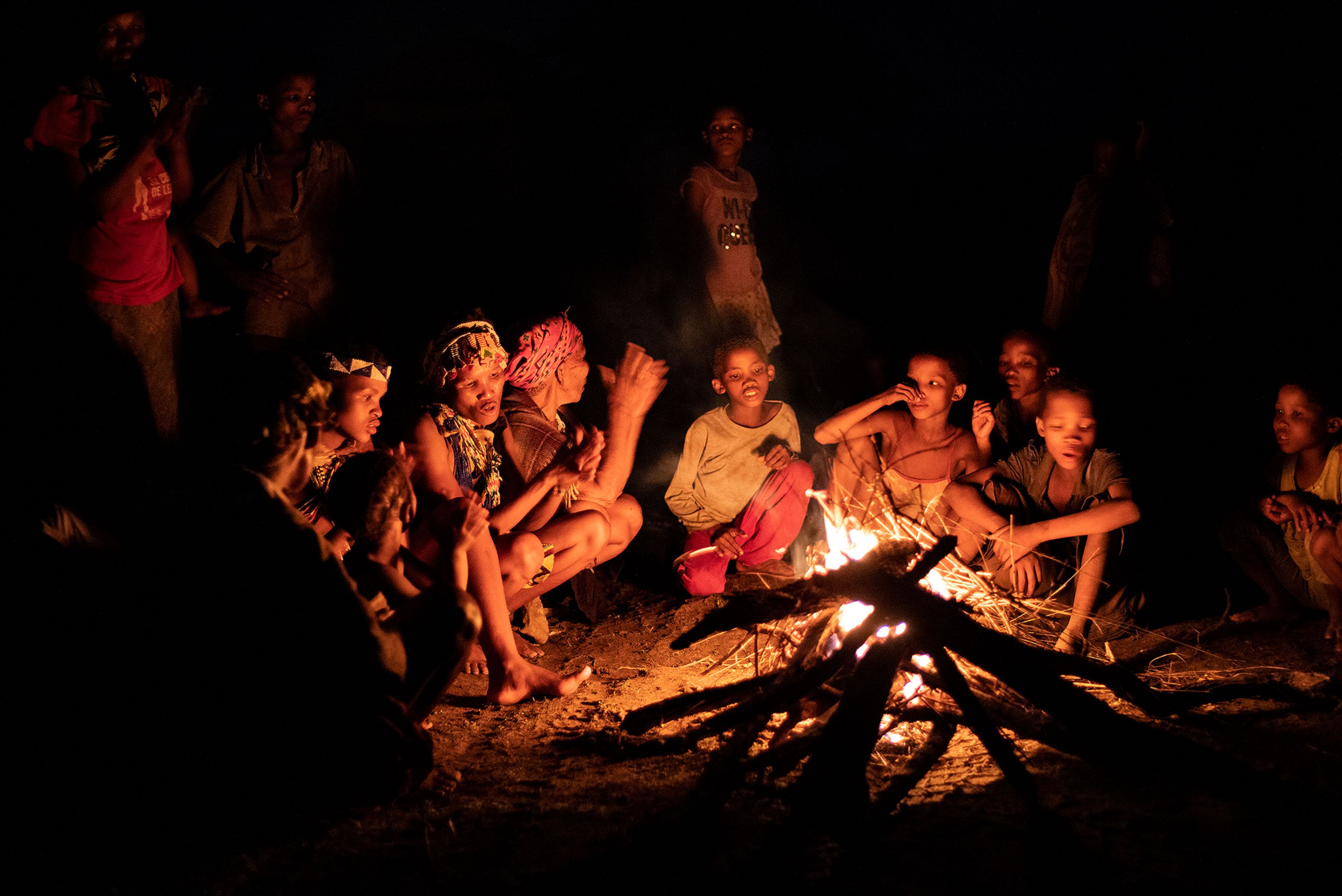 Der Stamm der San sitzt in der Nacht ums Lagerfeuer und singt traditionelle Lieder. Die Menschen schauen in das Feuer. Die älteren tragen traditionelle Kleidung des Stammes, die jüngeren tragen von Touristen mitgebrachte westliche Kleidung.